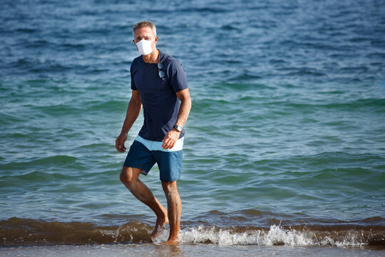 Uomo cinquantenne con mascherina protettiva e t-shirt blu sta in spiaggia e passeggia sereno di fronte al mare