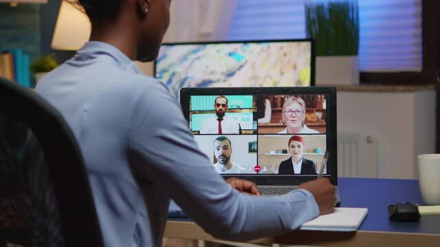 African freelancer working remotely discussing with partners online using laptop sitting in Livin room. Using modern technology network wireless talking on virtual meeting at midnight doing overtime