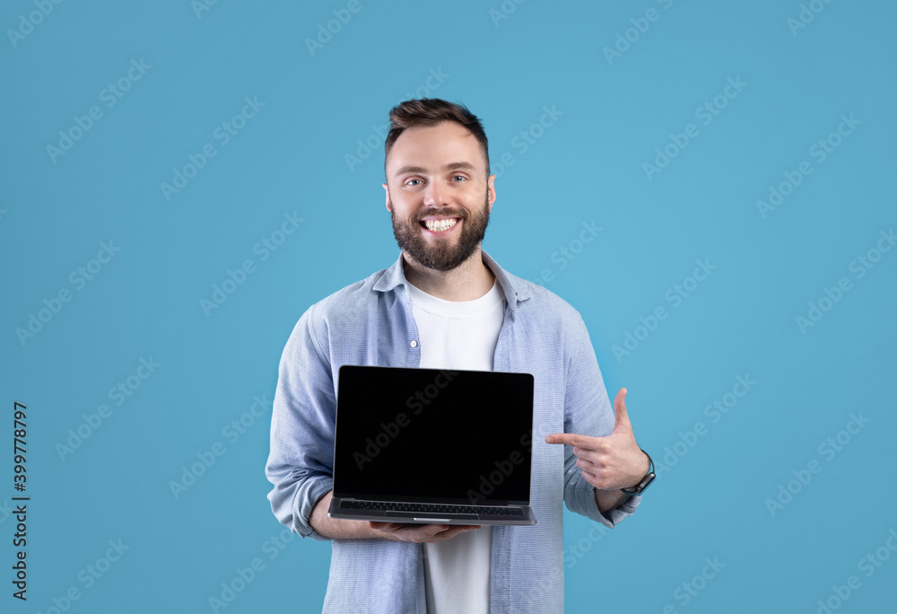 Poster portrait of happy bearded man holding laptop computer with empty screen over blue studio background,