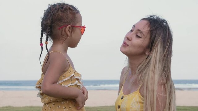 Tilt up of young Caucasian woman wearing yellow dress sitting on grass on the beach and talking to her little daughter. Then Caucasian boy suddenly hugging the woman