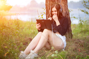 young beautiful woman watching movie on tablet with headphones in park under tree