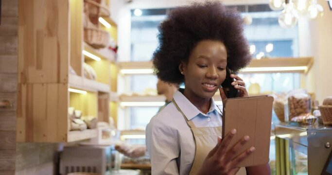 Close up. African American beautiful happy woman in apron calling and speaking on mobile phone using tablet while standing in bakery shop. Her coworker working on background. Bakehouse concept
