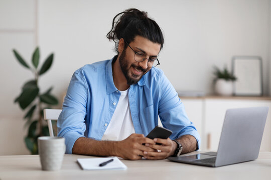 Eastern freelancer guy sitting at desk at home office and using smartphone