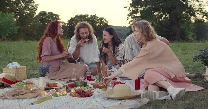 Millennial people eatting and laughing while sitting on grass and having picnic in park. Diverse friends spending good time together. Concept of leisure and friendship