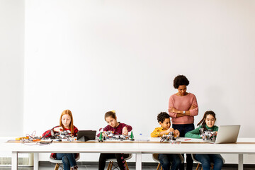 Happy kids with their African American female science teacher with laptop programming electric toys and robots at robotics classroom