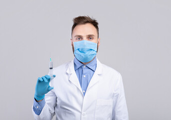 Immunization. Young male doctor wearing protection, holding syringe with coronavirus vaccine over grey background