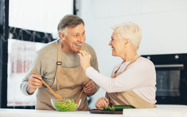 Senior Couple Feeding Each Other And Laughing Enjoying Cooking Indoors