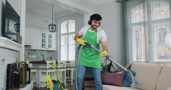 Portrait of likable joyful professional young cleaner in uniform which listening music in headphones while vacuuming sofa dust mites during cleaning up client's flat