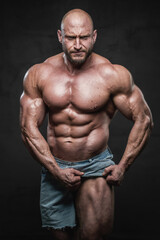 Fototapeta na wymiar Strong and shirtless bodybuilder with beard and bald head poses in dark background pulling his shorts and showing his muscular leg looking at camera with angry face.