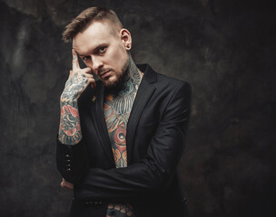 Portrait of a hipster person with tattooed body dressed in luxurious black jacket poses in dark background looking at camera with serious face.