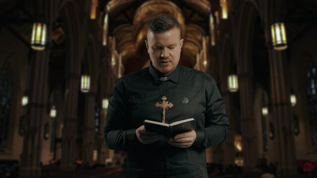 Closeup portrait of a young catholic praying priest on the background of the church.Priest holding is dressing in black, holding a crucifix and reading New Testament book. Low-key image.