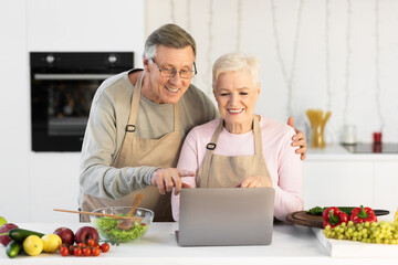 Senior Couple Cooking And Using Laptop Preparing Dinner In Kitchen