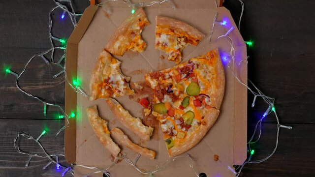 Top view - wooden christmas table with leftovers slices pizza in cardboard box. Christmas garlands decorations. After party concept.