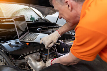 Fototapeta na wymiar Professional male mechanic worker using laptop under hood of car at the repair garage. Male car mechanic working using wrench tool for examining, repair and maintenance at car service shop