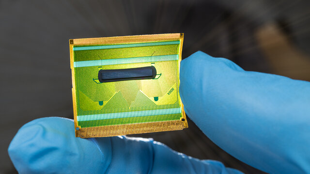 Close-up of flex circuit board in human fingers detail on dark background. Hand in blue glove holding green plastic flexible PCB for parallel transmission of digital data signals in LCD devices. Tech.