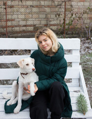 cute smiling teenage girl walks with her pet, young white dog in cold season. Relaxing sitting on a wooden bench outdoors. Active lifestyle, walks, joy every day
