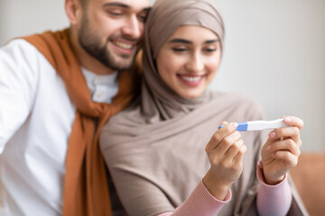 Joyful Muslim Couple Holding Pregnancy Test Sitting Together Indoors