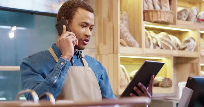Close up of joyful African American young handsome man worker in apron stands in bakery shop, speking on smartphone and using tablet device. Business concept. Retail industry