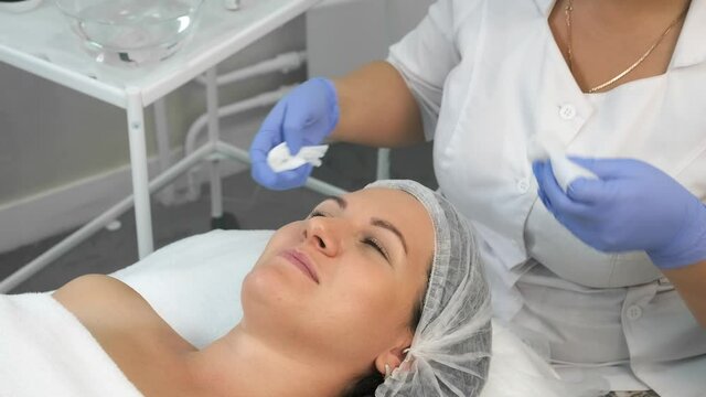 Cosmetologist in gloves is wiping cleaning woman's face using napkins before peeling procedure. Doctor is making beauty procedure in cosmetology clinic. Care about patient's face skin.