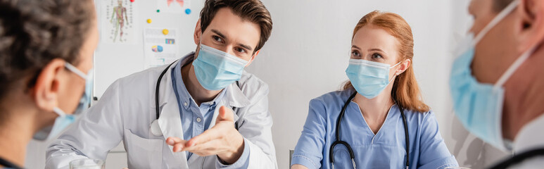 Doctor in medical mask sitting near colleagues and pointing with hand at african american nurse in...