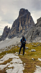 Fototapeta na wymiar Man with big backpack and sticks, hiking in high Italian Dolomites. There are many sharp peaks in front of him. He is standing on a big boulder. Lots of lose stones and landslides. Sunny day. Outdoor