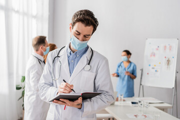Doctor in medical mask writing in notebook with blurred colleagues working on background