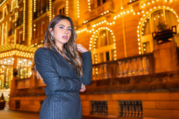Christmas shooting, a Caucasian brunette girl in the city at night by the Christmas lights. Elegant black christmas or new year's eve dress with black boots