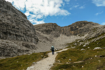 Fototapeta na wymiar A woman hiking in a high and desolated mountains in Italian Dolomites. The lower parts of the mountains are overgrown with moss and grass. Raw and unspoiled landscape. Clear and sunny day. Solitude