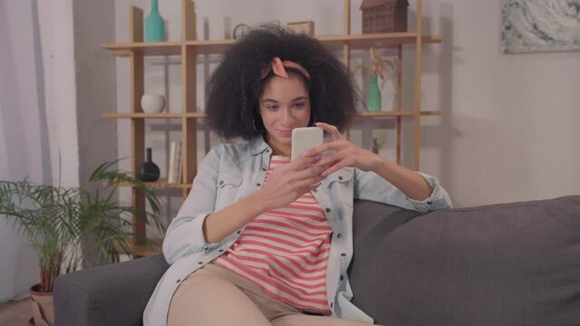 young hispanic woman messaging on smartphone and sitting on couch