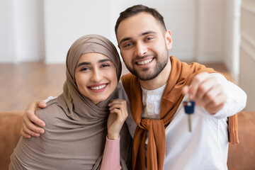 Cheerful Muslim Young Family Showing New House Key Sitting Indoors