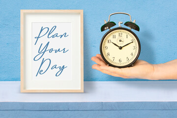 Hand holding an alarm clock next to a wooden frame with the sentence "Plan your time" as time management concept.