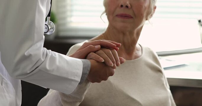 Practitioner doctor wear white coat holding hand of older patient during talk at hospital, close up. Reliable therapist provide psychological support to aged client of clinic, showing empathy concept