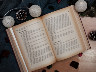 Opened old book pages with Christmas lights decorations and scarf on wooden surface with pine cone,...