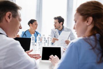 Doctor holding paper near african american nurse and colleagues with devices on blurred foreground