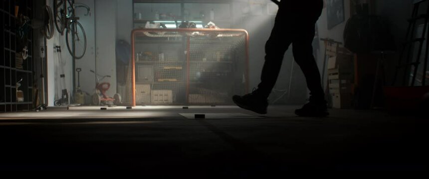 Silhouette of Caucasian male hockey player practicing slap shots inside his garage. Shot with 2x anamorphic lens