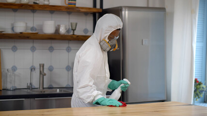 Disinfector in protective suit and mask spraying detergent and wiping table in house kitchen