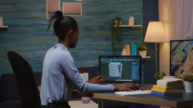 African lady enginer analysing digital mechanism of container checking prototype of project looking on laptop sitting at desk in living room office late night. Black employee using modern technology