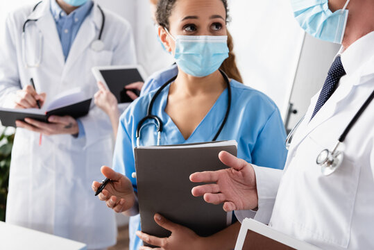 African american nurse in medical mask talking to doctor with blurred colleagues on background