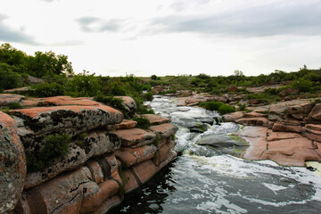Waterfall flows among high rocks, red stones and cliffs, green bushes and trees
