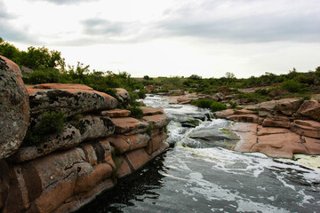 Waterfall flows among high rocks, red stones and cliffs, green bushes and trees