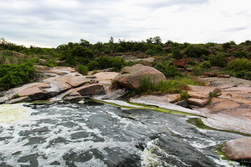 Waterfall flows among high rocks, red stones and cliffs, green bushes and trees