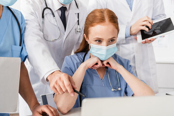 Redhead nurse in medical mask looking at laptop near multicultural colleagues standing behind on blurred foreground