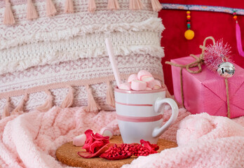 Obraz na płótnie Canvas Close-up view of a pink home corner with a gift package on a blanket, a mug with hot cocoa with marshmallows and Christmas decorations. Holidays and happy people concept
