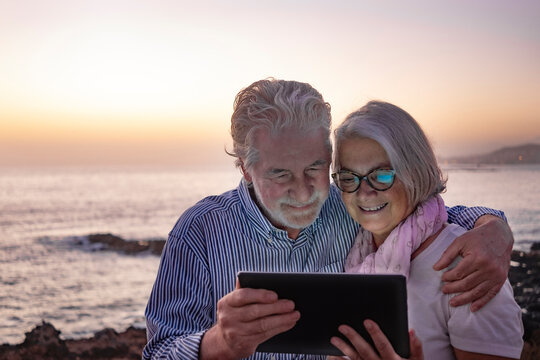 Embraced attractive senior couple white-haired looking at digital tablet in the dusk sitting on the beach with horizon over water on background. Happy retirement concept together