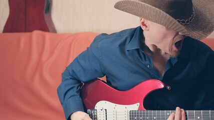 Caucasian man in cowboy hat plays electric guitar. Man shouts loudly, aggressively. Interior looks...