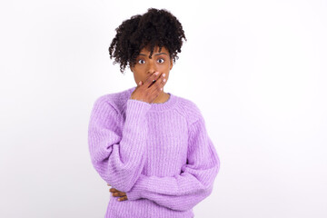 Emotional African American woman wearing purple knitted sweater against white wall gasps from astonishment, covers opened mouth with palm, looks shocked at camera. Reaction concept
