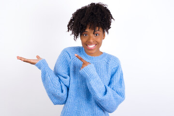 oung beautiful African American woman wearing blue knitted sweater against white wall pointing and...
