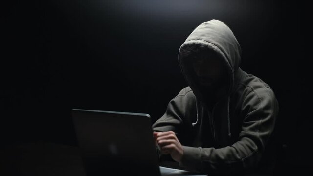 Remote view of unrecognizable hacker man stretching hands and starting to typing on laptop computer keyboard breaking password. Hooded bearded freelancer programmer hacking online site or password.