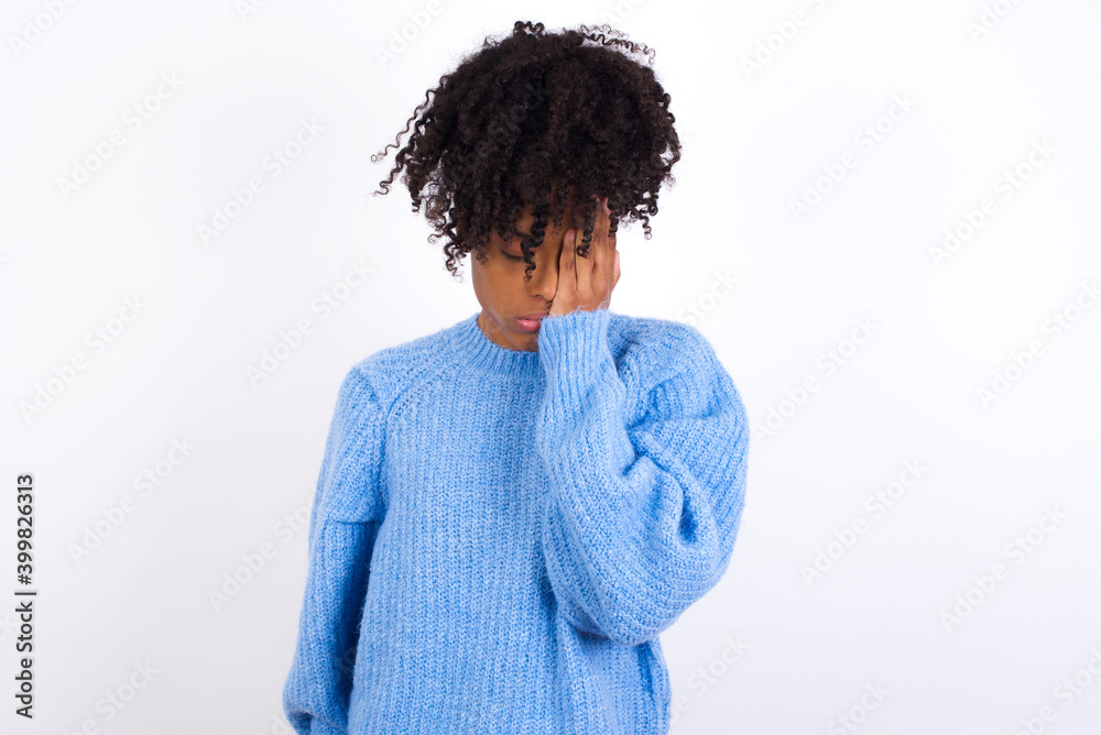 Wall mural Tired overworked oung beautiful African American woman wearing blue knitted sweater has sleepy expression, gloomy look, covers face with hand, has eyes shut, gasps from tiredness, fatigue after party