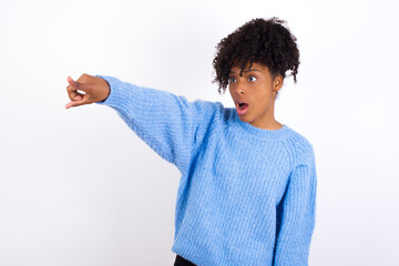 Young beautiful African American woman wearing blue knitted sweater against white wall Pointing with finger surprised ahead, open mouth amazed expression, something on the front.